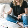 Woman wearing two gold necklaces reading Bible while drinking coffee